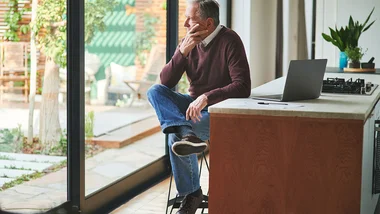 Middle-aged man sitting at a kitchen island with his laptop, looking inquisitively out a window 