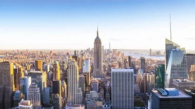 This image shows the skyline of New York City, featuring the Empire State Building prominently in the center. The cityscape includes various skyscrapers and buildings, with a clear sky above, suggesting it is a bright day. The background shows the river and distant landscape.